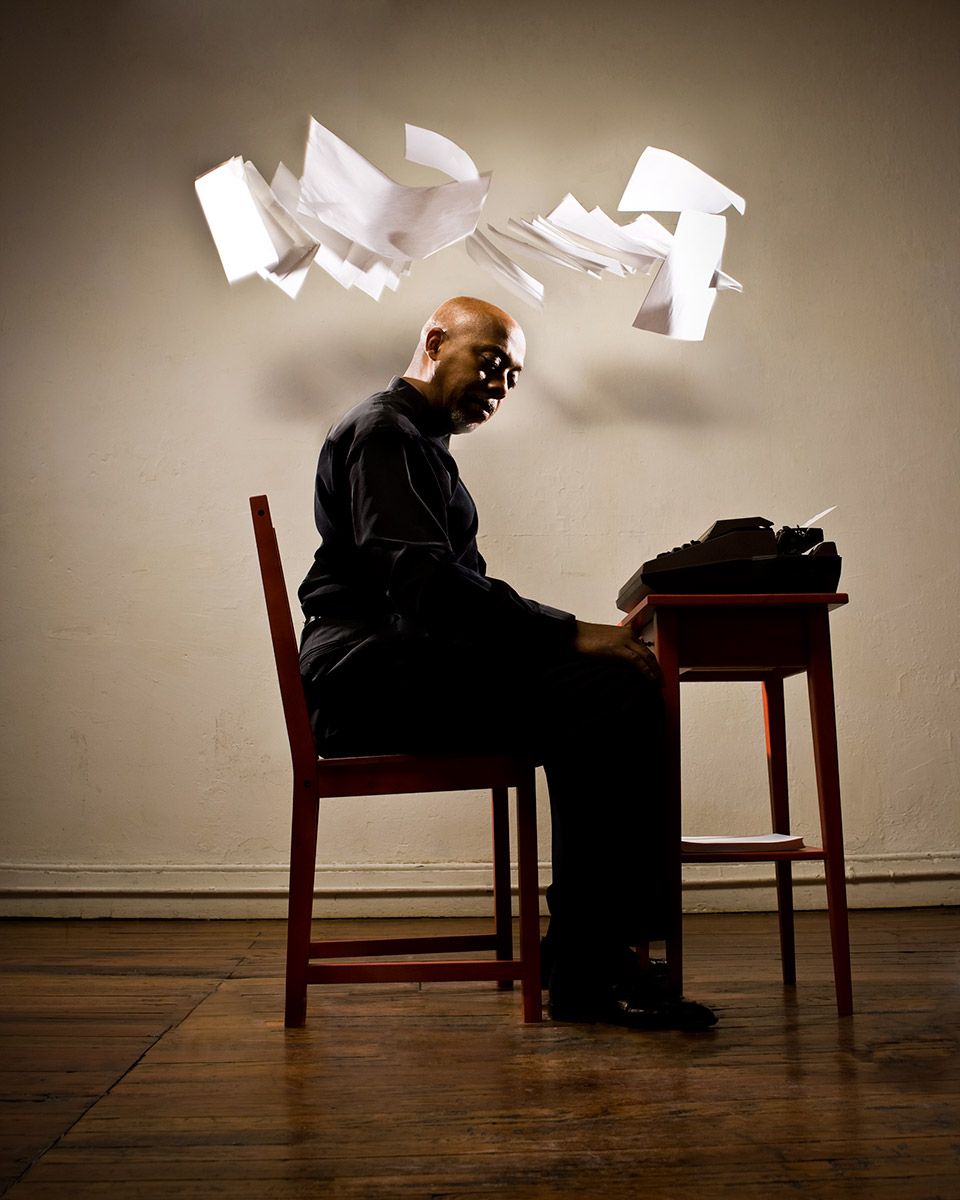 Man sitting at typewriter with papers flying above his head in dramatic studio shot.