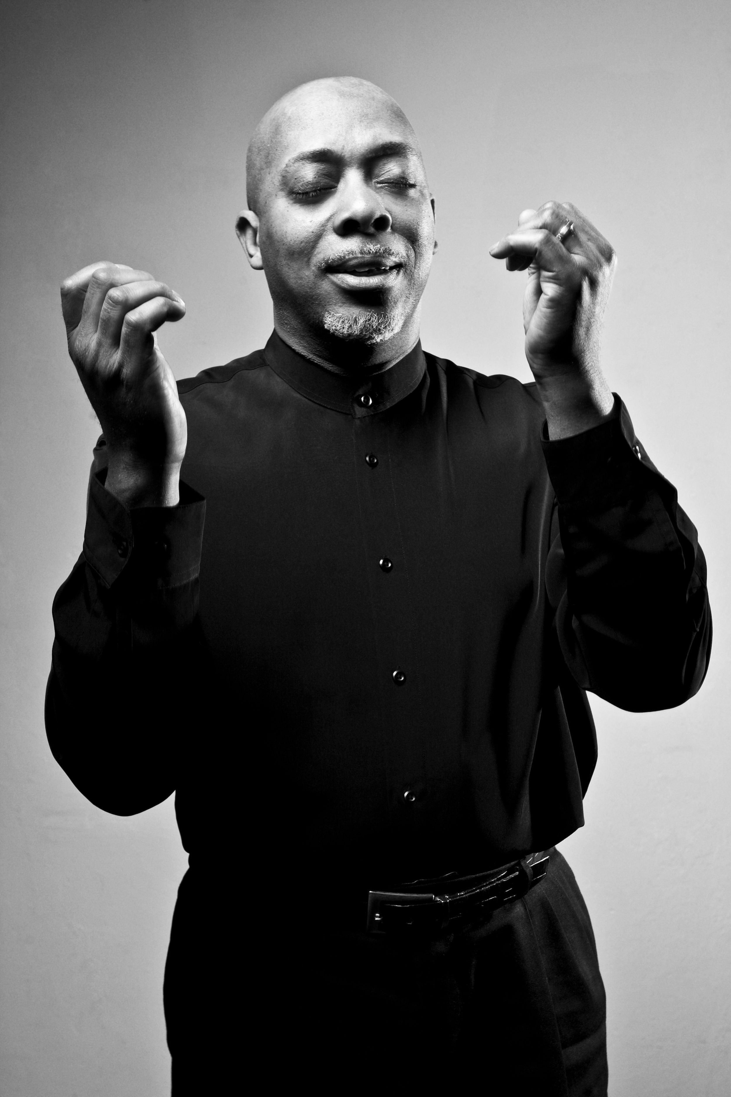 Bald Black man in black shirt performing with eyes closed and hands raised in expressive gesture.