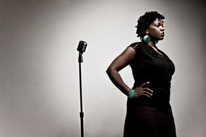 Black woman in elegant black dress standing confidently beside vintage microphone stand.