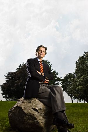 Businessman in suit sitting on large rock with arms crossed in green park setting.