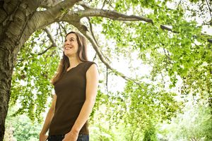 Smiling woman in brown sleeveless top standing under lush green tree in sunlight.