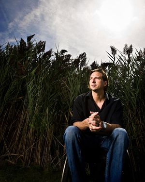 Young man sitting outdoors among tall reeds gazing upward at dramatic cloudy sky.
