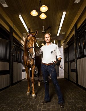 Man in white polo shirt leading chestnut horse through elegant stable corridor.