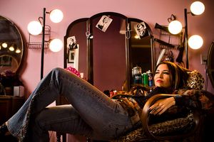 Woman in jeans reclining in chair beside vintage vanity mirror with round lights.