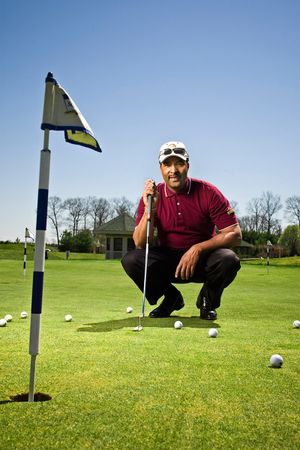 Golfer crouching on green near flag with golf balls scattered around on sunny day.