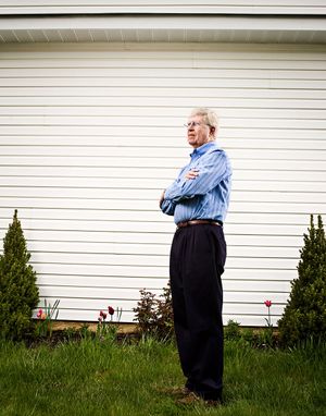 Older man standing with arms crossed beside white building with small shrubs and tulips.