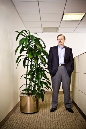 Businessman in suit standing beside tall indoor plant in office corridor.