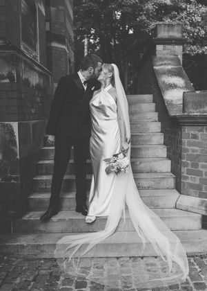 Black and white photo of a bride and groom sharing a kiss on stone steps outside a historic building.