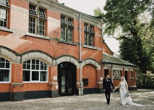 Bride and groom walking hand in hand outside a charming red brick wedding venue surrounded by lush trees.