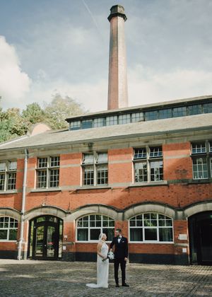 Bride and groom stand before a historic red brick mill building with tall chimney stack on their wedding day.