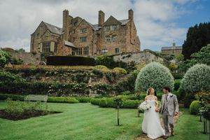 Bride and groom walking in manicured gardens of a grand stone manor house on their wedding day.