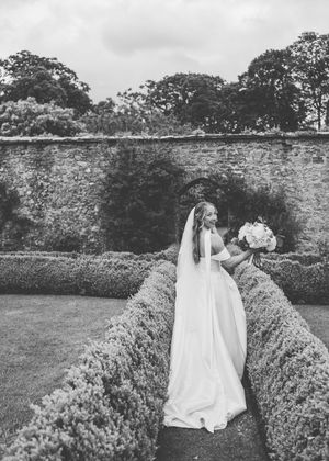 Bride in white gown and veil holds bouquet in a formal garden with stone wall backdrop, black and white photo.