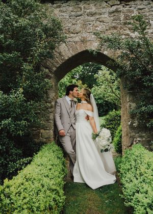 Bride and groom kissing under a stone arch surrounded by lush greenery, bride holding white bouquet in elegant wedding attire.