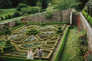 Bride and groom walking hand in hand through an ornate formal garden with geometric topiary hedges and stone walls.