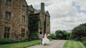 Bride and groom dancing outside a historic stone manor house surrounded by lush green gardens on a cloudy day.
