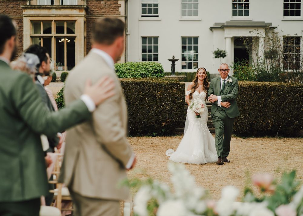 Bride walks with father toward groom during outdoor wedding ceremony at elegant English manor house.