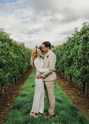 A romantic couple embraces in a lush vineyard row with green vines and cloudy skies overhead.