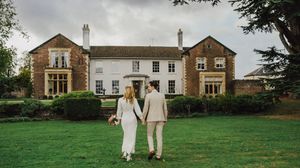 A couple walks hand in hand across a lush green lawn with historic brick buildings in the background.