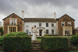 A wedding couple poses in front of a rustic stone building with white trim and lush greenery.