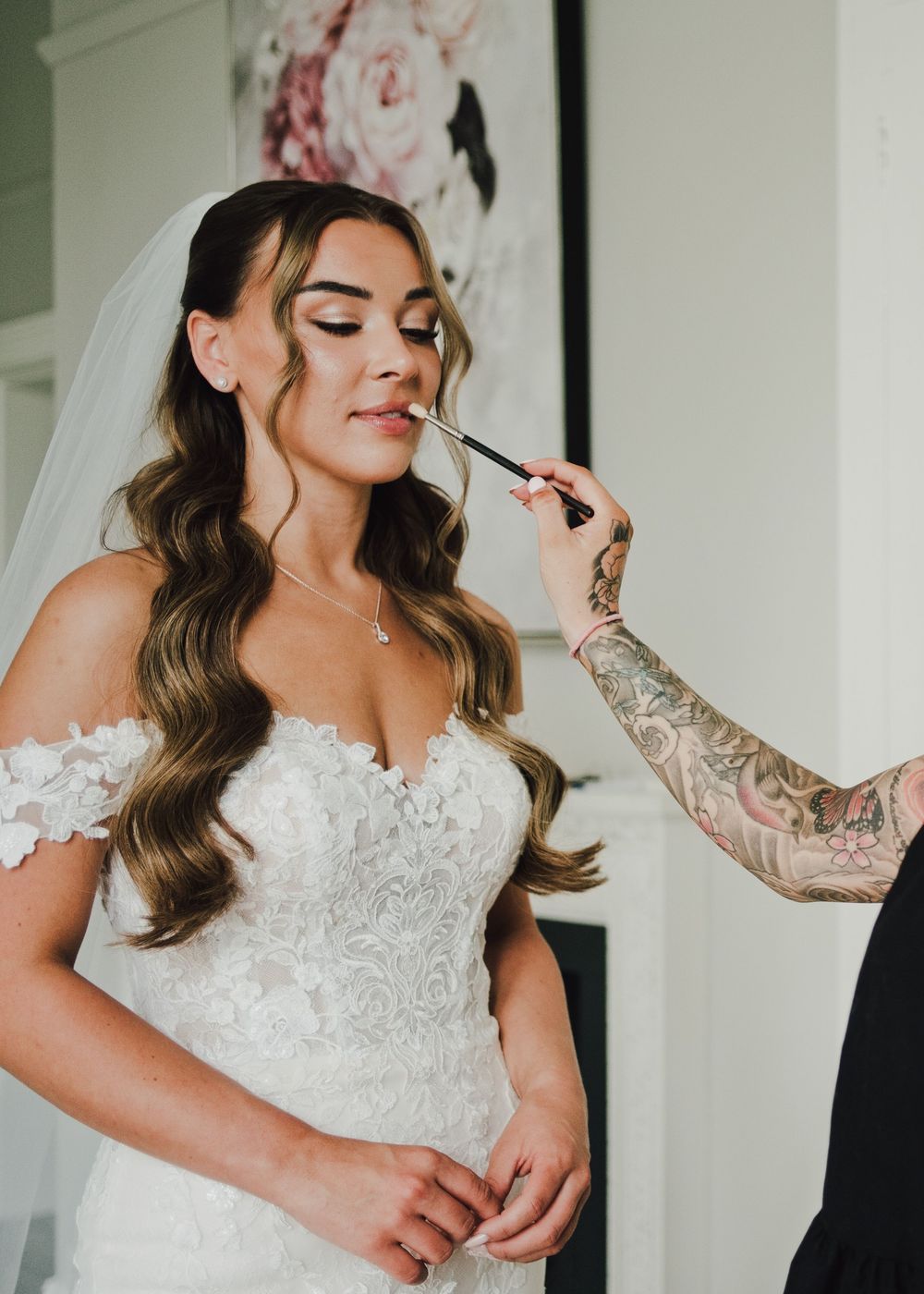 A bride in a lace off-shoulder gown gets lipstick applied by a tattooed makeup artist during wedding preparations.