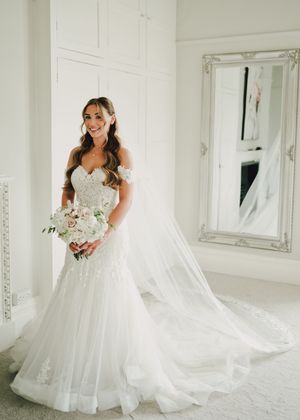 Bride in off-shoulder lace mermaid wedding gown holding white bouquet, standing in elegant white room with ornate mirror.