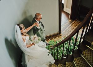 Bride in white gown holds bouquet as older man kisses her hand on elegant curved staircase.