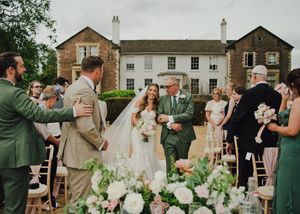 Bride walks down outdoor aisle with father at elegant stone manor wedding ceremony, guests in sage green attire.