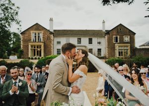 Bride and groom share a first kiss at outdoor wedding ceremony in front of elegant stone manor house.