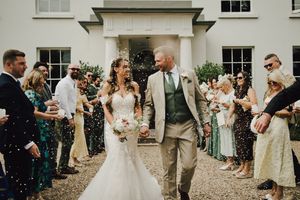 Bride and groom walk through confetti shower outside elegant white manor house surrounded by cheering wedding guests.