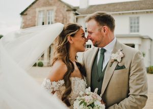 Bride and groom share a romantic moment, touching foreheads, with veil flowing and a floral bouquet outside an elegant venue.