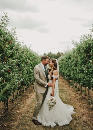 Bride and groom share a romantic kiss in a lush apple orchard, bride in white gown, groom in tan suit.