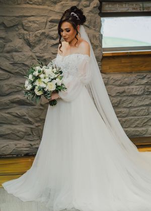 A bride in a flowing white wedding gown and veil poses against a rustic stone wall background.