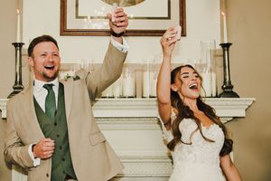 Joyful newlyweds celebrate at wedding reception, groom in tan suit raises glass beside bride in white gown near fireplace.