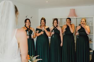 Bridesmaids in matching emerald green gowns react with surprised expressions while holding champagne glasses.