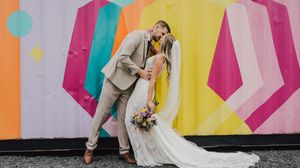 A couple poses together against a vibrant geometric mural with pink and yellow angular shapes.