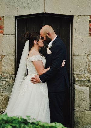 Bride and groom share a romantic forehead touch in wedding attire before a black door at a stone venue.