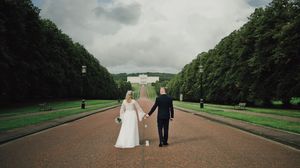 A bride and groom hold hands walking toward a grand white building along a tree-lined path on a cloudy day.