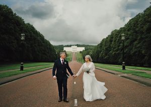 Bride and groom hold hands on a grand tree-lined path at Stormont Estate, Belfast, on their wedding day.