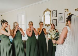 A group wearing sage green bridesmaid dresses stands together in an interior space while getting ready for a wedding.