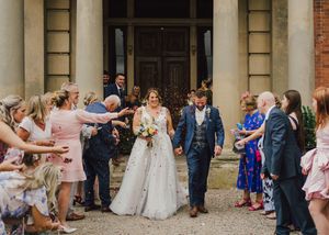 Wedding guests celebrate by throwing confetti as newlyweds exit a historic brick building with stone columns.