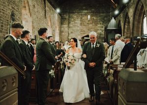 Bride walks down church aisle with father in stone chapel, groom and groomsmen in dark suits await at altar.
