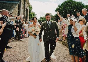 Bride and groom walk through confetti shower at outdoor wedding ceremony, guests lining path at rustic stone venue.