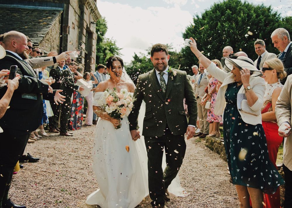 Bride and groom walk through confetti shower at outdoor wedding ceremony, guests lining path at rustic stone venue.