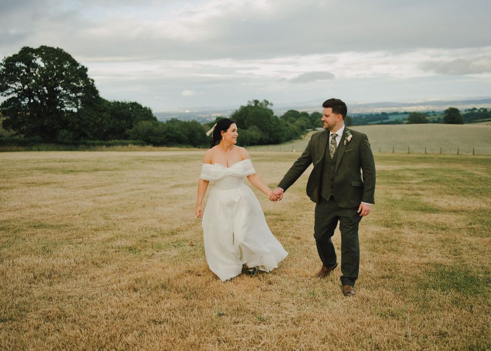 Bride and groom holding hands walking through a green field on their wedding day in the English countryside.