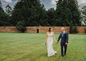 A couple holding hands walks across a green lawn beside a stone wall with trees in the background.