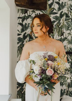 Bride in off-shoulder gown holding colorful bouquet against floral wallpaper.