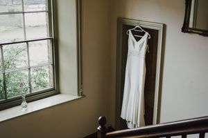 A sleek white wedding dress hangs on a wooden door frame near a window in a minimalist room.