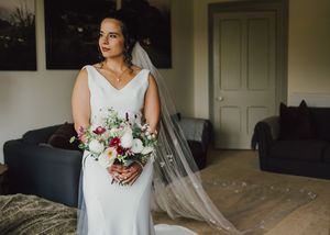 A bride in a white fitted wedding dress holds a pink and white floral bouquet while standing in a room.