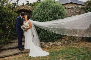 A wedding couple standing by a stone wall as a long white veil billows in the wind during an outdoor ceremony.