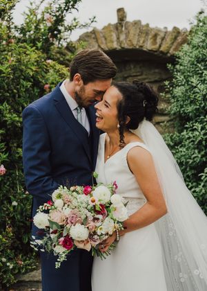 Newly married couple share an intimate moment in a garden with stone archway while holding a pink and white bouquet.
